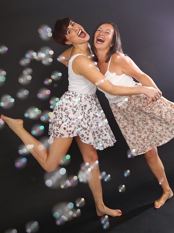 Zwei Mädchen lachend in Kleidern vor Seifenblasen im Gegenlicht.